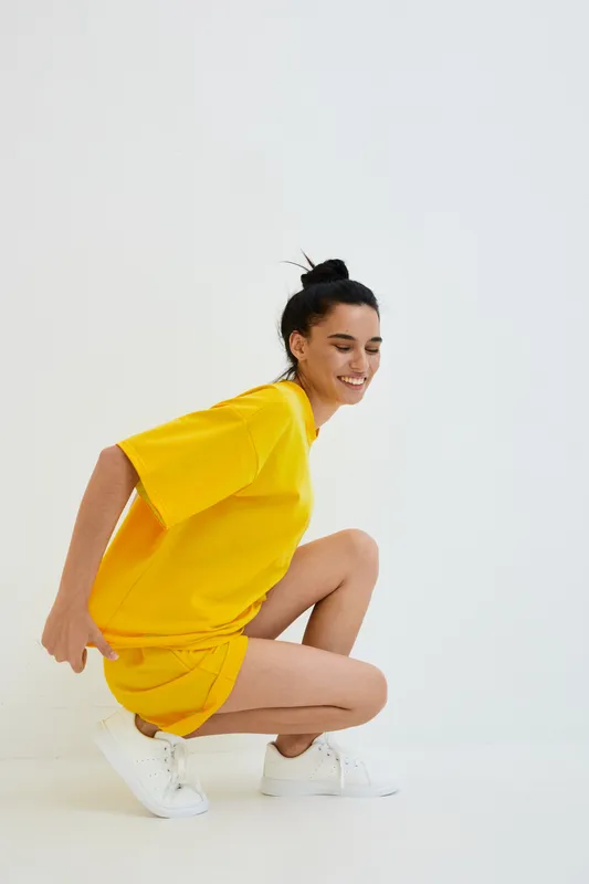 smiling woman in yellow casual outfit crouching on white background, youthful, joyful, relaxed mood, minimalistic studio environment, fresh summer style