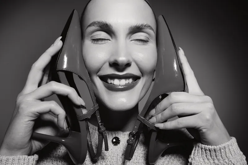 Happy Caucasian woman with light skin holds high heel shoes by her face smiling with closed eyes in black and white. Concept for fashion, joy, and footwear lifestyle.