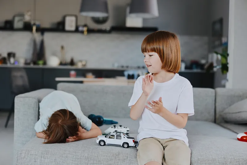 Playful boy sitting on gray sofa, engaging with a toy car, while another child leans forward in a cozy living room. Bright and inviting atmosphere enhances childhood joy.