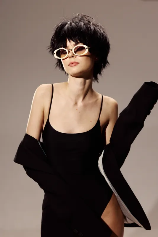 Stylish woman with short hair wearing sunglasses and a black outfit, posing confidently against a plain background.