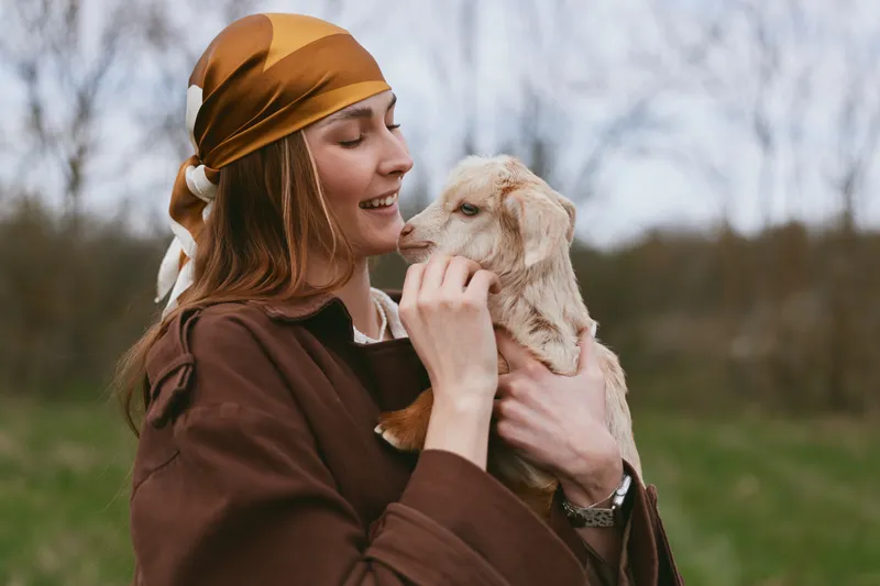 Happy woman outdoors, holding a goat kid close, warm sunlight, soft breeze, casual attire, gentle bond, natural colors, countryside landscape, candid moment, peaceful atmosphere