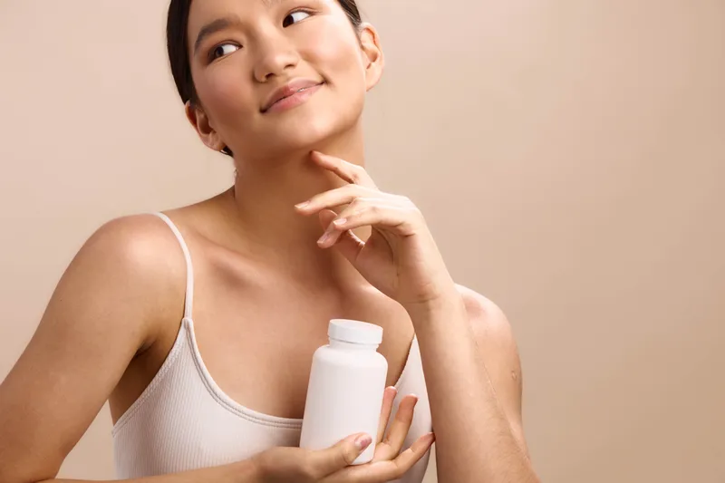 Asian woman smiles softly while holding a bottle in a light neutral setting, presenting a calm skincare routine and healthy beauty vibe for a soothing professional portfolio