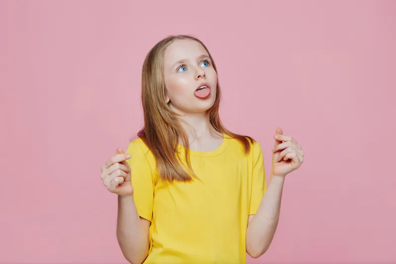 A cheerful girl with long blonde hair wearing a yellow shirt, playfully sticking out her tongue against a pink background, showcasing a fun and carefree expression