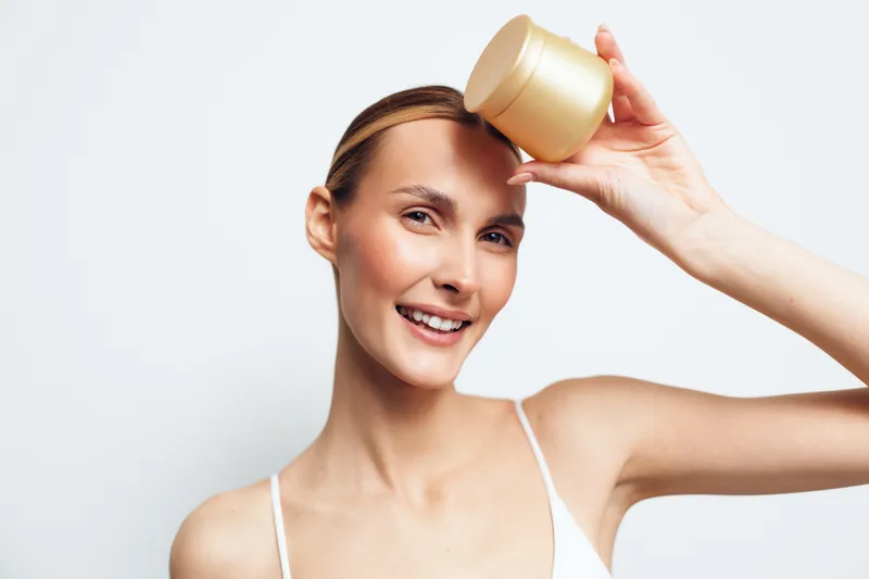 Young woman with brunette hair smiles as she applies a cosmetic cream from a small jar to her scalp in a bright studio, presenting a relaxed confident moment, natural light