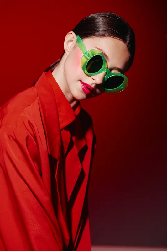 Elegant woman wearing bold green sunglasses and bright red coat poses against a red background. Creative unique beauty portrait combining fashion and colorful style concepts.