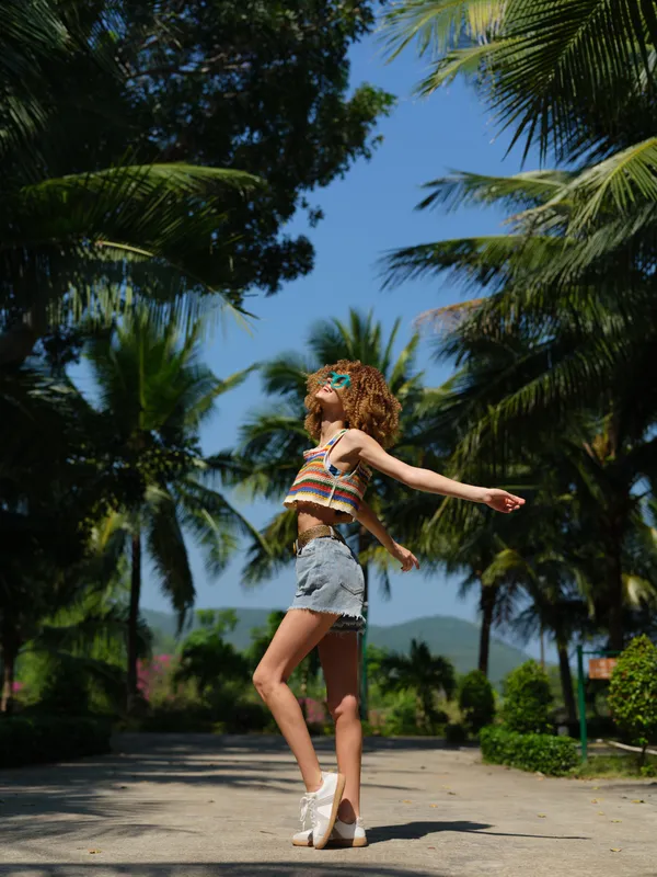 young woman with curly hair wearing colorful top and denim shorts standing with arms outstretched on sunny tropical path, surrounded by palm trees and clear blue sky