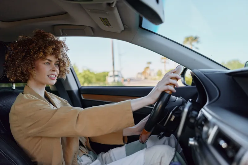 Smiling woman with curly hair driving a car, interior view highlighting steering and dashboard, casual fashion and relaxed lifestyle under warm retro lighting. Soft glow and daylight set mood for