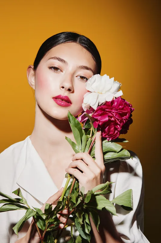 Young woman with glossy pink lips and soft makeup holds light and dark flowers close on mustard background. Style beauty concept with floral fashion and creative mood.