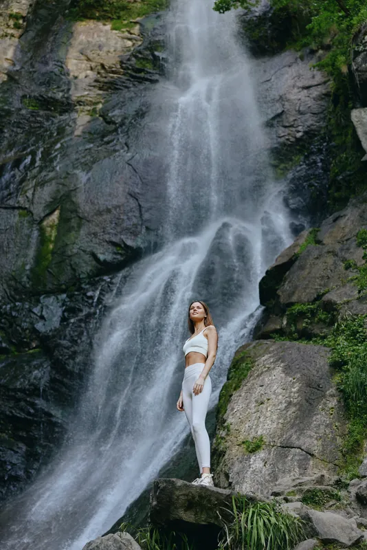 Woman standing near large waterfall in white outfit, surrounded by rocks and greenery, displaying calm and peaceful outdoor nature scene.