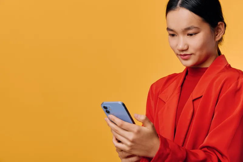 Young woman in red blazer holding and looking at smartphone with slight smile, isolated on orange background. Technology, communication, casual, modern, portrait with mobile device.