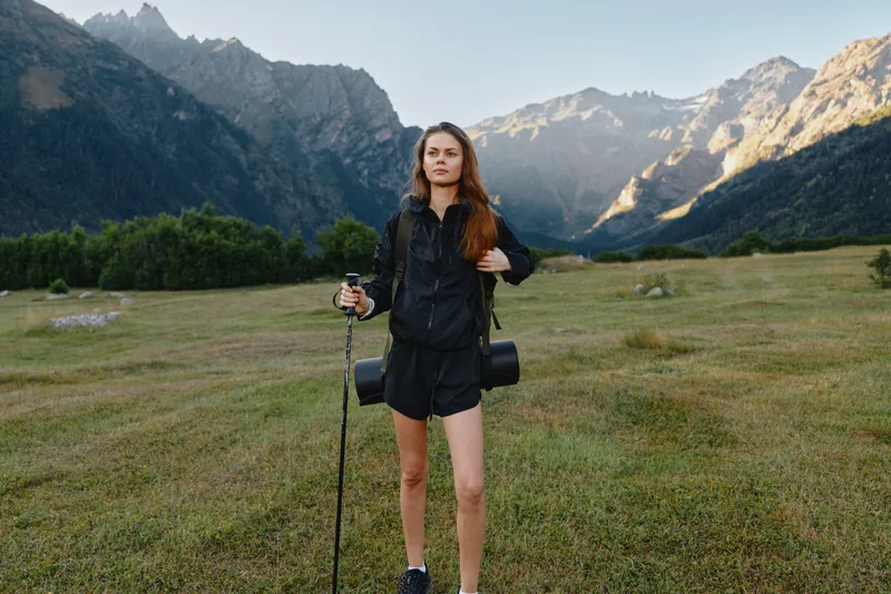 A solo female hiker stands in a wide meadow with a trekking pole and backpack, set against dramatic mountains. She gazes ahead with confidence, inviting outdoor adventure.