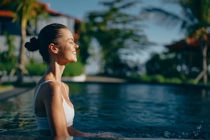 Smiling woman in a white swimsuit enjoys a sunny poolside moment, hair in a neat bun. Bright outdoor light highlights a relaxed lifestyle, wellness, and self care beside clear water on a tropical