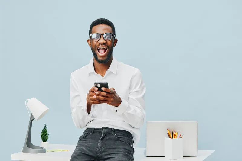 Smiling man in white shirt using mobile phone at desk with modern lamp and stationery on light blue background, joyful expression, casual office attire, professional workplace environment, happiness, 