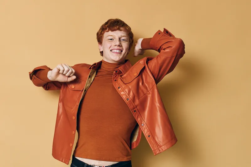 Young man with red hair wearing orange leather jacket and sweater posing and smiling on beige background. Casual style, studio portrait, happy expression and confident look outdoors.