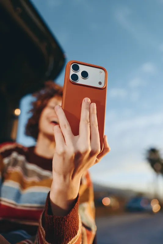 Woman lifestyle outdoors selfie with smartphone, holding phone close to face, hand gesture near lens, soft blurred street and sky background, warm evening light, casual portrait moment