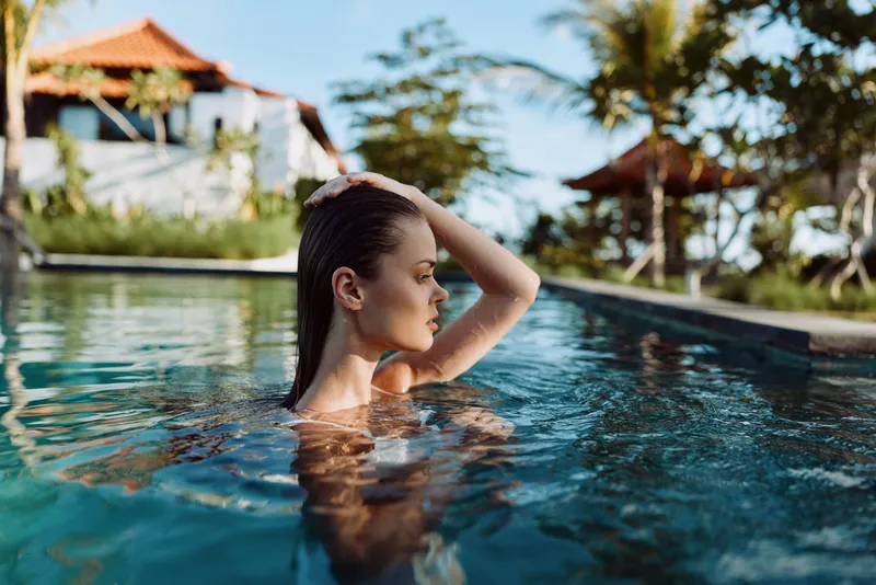 A relaxed woman in a bright pool raises her hand to her head, water gliding over her shoulders. A tropical resort backdrop creates a calm lifestyle moment focused on leisure and escape.