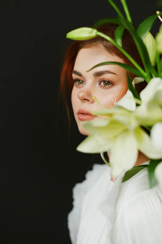 A serene portrait of a young woman with brown hair, wearing a white outfit, surrounded by white lilies against a dark background, conveying elegance and beauty