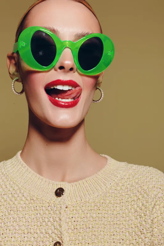 Young woman wearing large green sunglasses and red lipstick smiles playfully with tongue out. She wears hoop earrings and a beige knitted sweater on a neutral background.
