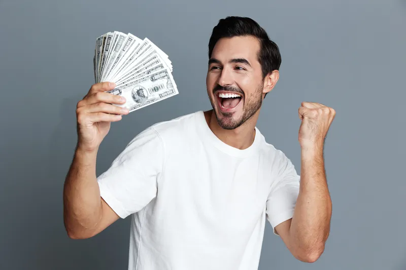 Excited smiling young man holding cash in hand celebrating success over gray background in studio portrait. Happiness and financial success concept.