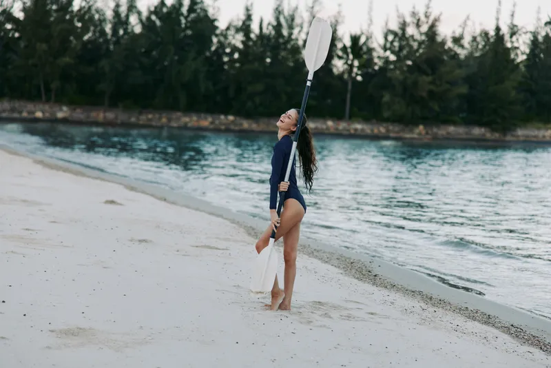 A female athlete stands on a bright sandy shore holding a paddle, wearing a dark swimsuit and long sleeves, as calm sea water and pine trees create a tranquil scene.
