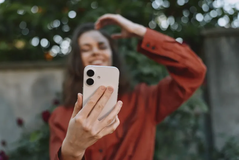 Smiling woman in a red jacket taking a selfie outdoors, with green foliage in the background. Lifestyle, casual beauty, self expression, warm natural lighting