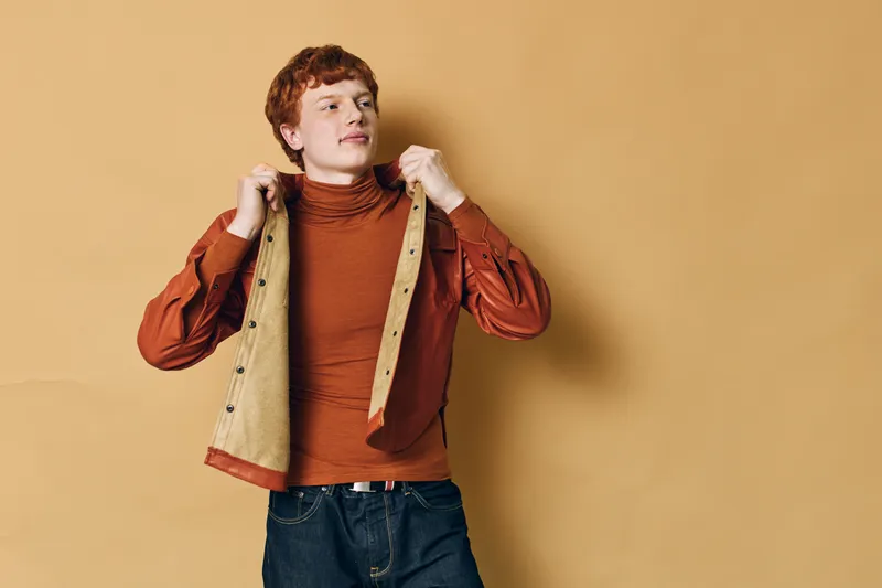Young man with red hair wearing orange turtleneck and beige jacket posing against beige background. Casual style, confident look, fashion and youth lifestyle portrait.