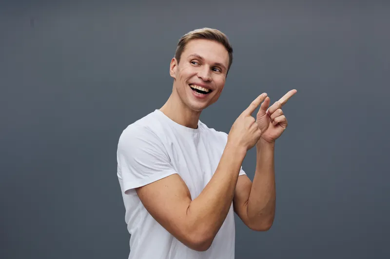Smiling young man with short light brown hair wearing a white t-shirt pointing sideways against a plain gray background. Relaxed, cheerful expression. Studio portrait. People lifestyle concept.