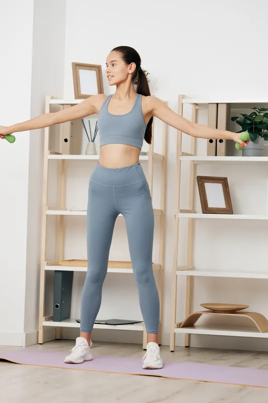 Woman fitness workout scene in a home gym with a calm, focused expression. She stands on a mat, arms extended holding light dumbbells, performing a lateral raise in athletic apparel within a bright,