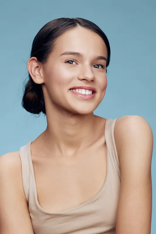 Smiling young woman with natural makeup and light clothing against a soft blue background, showcasing beauty, confidence, and a positive mood