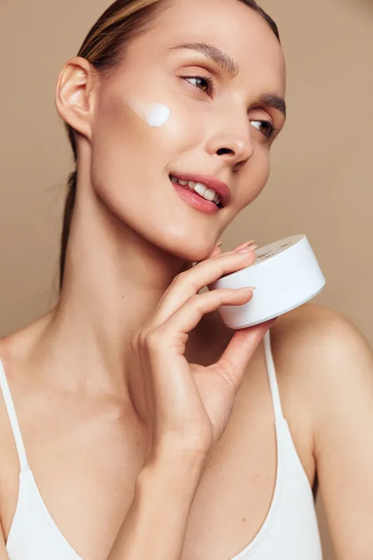 Young woman with clear skin applying facial cream on cheek, holding white jar, natural makeup, soft lighting, skincare routine, closeup portrait, beige background.
