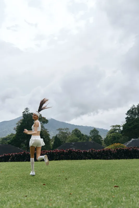 Young female athlete running on a green field with a joyful expression, wearing sporty white attire against a cloudy backdrop, capturing the essence of an active lifestyle
