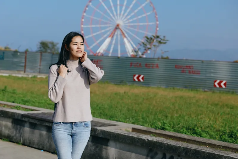 Woman enjoying a sunny day outdoors near a ferris wheel, wearing a light sweater and jeans, gazing into the distance in a spacious park setting.