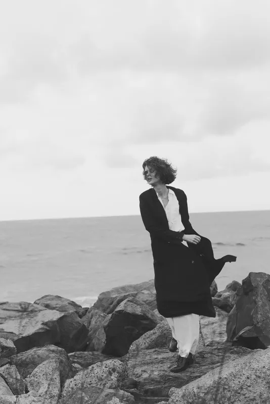 Woman in a dark coat stands on rugged rocks by the sea, wind tousling her hair, a monochrome scene that conveys solitude, resilience and a moody coastal atmosphere