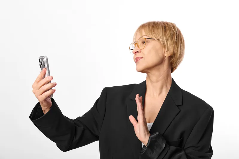 Woman in business suit holds smartphone, stop refusal gesture for communication, isolated on white background, mature female with glasses, modern technology caution