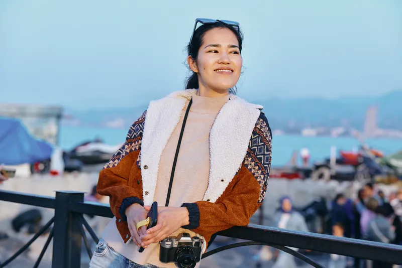 Woman with camera enjoying outdoor beach scene, casual fashion and travel vibe, smiling amid sun, sea and coast, capturing candid moment by boardwalk and breeze