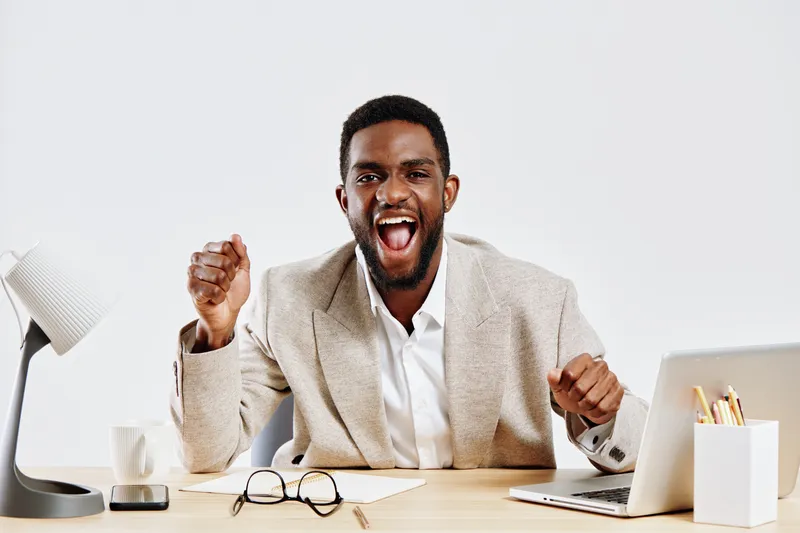 Happy young African American man in beige blazer celebrates success at office desk, smiling and raising fist, surrounded by office supplies, cheerful mood, professional attire, upbeat atmosphere, stud