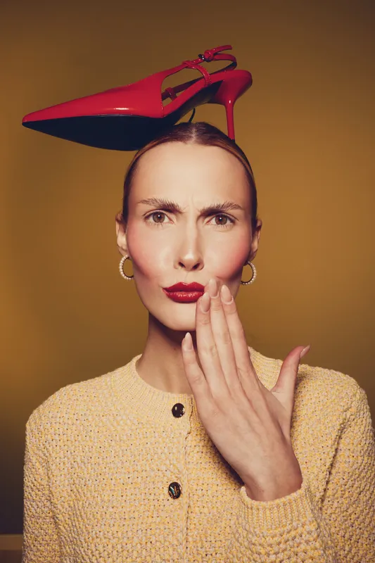 Woman with red lipstick and hoop earrings holds hand near mouth with red high heel shoe balanced on head. Concept of fashion, style, and playful attitude.