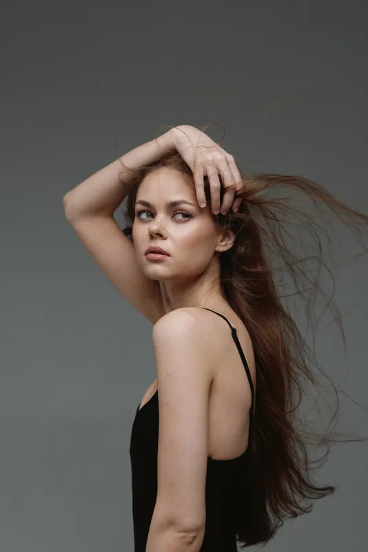 Portrait of a young woman with long hair wearing a black dress posing thoughtfully against a gray background