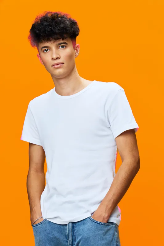 young man wearing a white t-shirt stands confidently against a vibrant orange background with hands in pockets, casual style and natural curly hair.