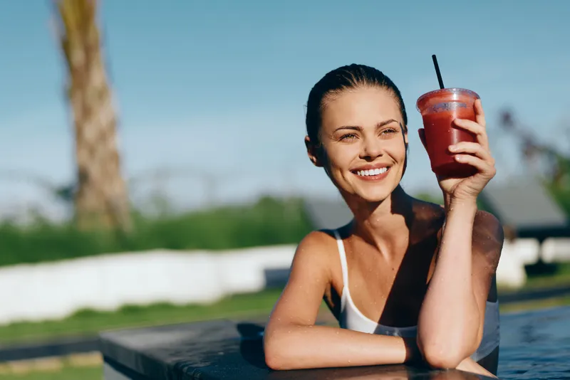 Cheerful woman outdoors, smile on her face, enjoying a refreshing drink by the pool, sunlit scene, casual pose, warm breeze, vibrant colors, carefree lifestyle