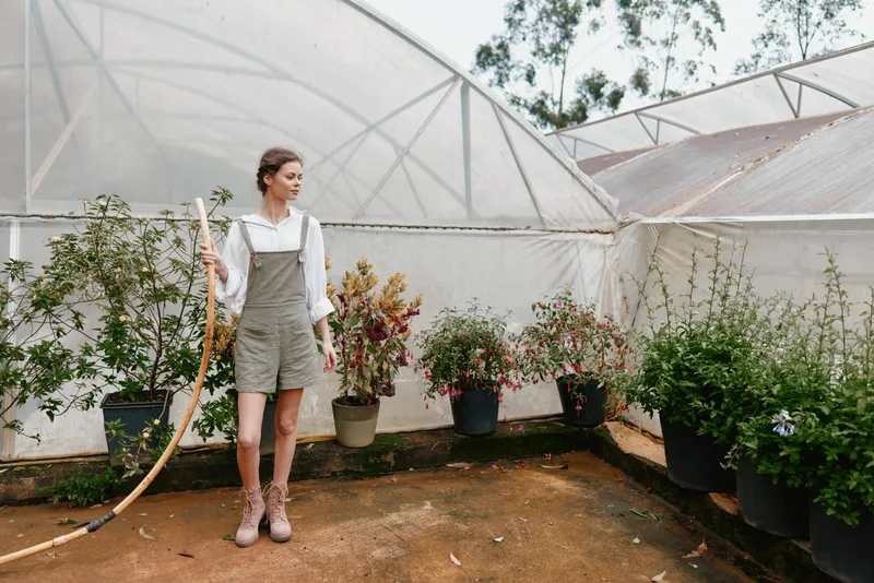 Young woman in a greenhouse wearing casual overalls and a white shirt, surrounded by lush plants, expressing tranquility and connection with nature