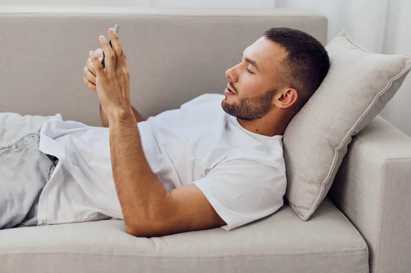Relaxed man using smartphone while lying on a couch, dressed casually in a t-shirt. The soft light creates a warm and cozy atmosphere, perfect for leisure moments.