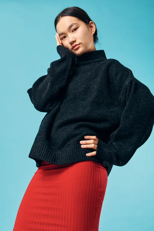 Young woman wearing oversized black sweater and red skirt standing against blue background with hand on face and thoughtful expression, fashion portrait with casual style.