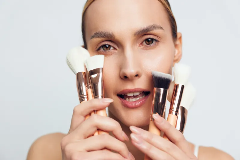 Young woman holding multiple makeup brushes close to her face with neutral expression on light background. Concept of beauty tools, cosmetics, and skincare routine essentials.