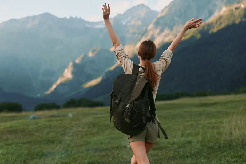 A woman with a backpack stands in a green meadow, arms raised toward distant mountains, capturing a moment of achievement, freedom, and outdoor adventure.