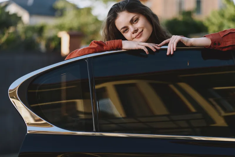Young woman with dark hair leans on a glossy car window outdoors, smiling softly in a relaxed pose as warm sunlight highlights her red shirt and a gentle breeze plays with her hair against an urban