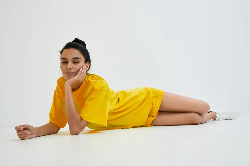 Relaxed woman in bright yellow casual outfit lying on white floor, wearing sneakers, with a calm expression and hair tied in a bun.