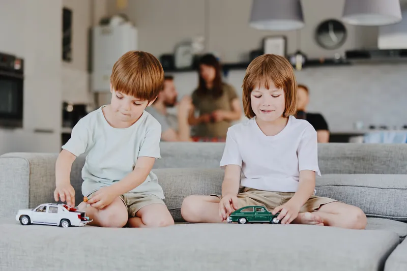 Two young boys playing with toy cars on a cozy sofa, showcasing joy and childhood innocence in a bright, modern living room setting.