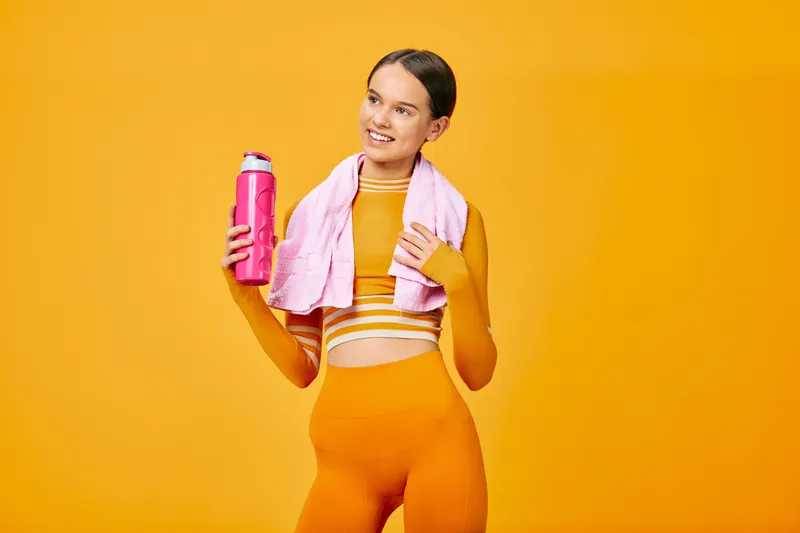 Woman in bright orange athletic outfit with a pink towel draped over shoulders holds a pink water bottle against a vivid orange background, showcasing fitness and active lifestyle.