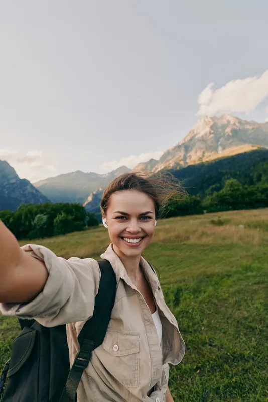 A woman on a daytime hike in a mountainous landscape smiles at the camera, wearing a light shirt and backpack. A lifestyle scene focused on outdoor exploration.